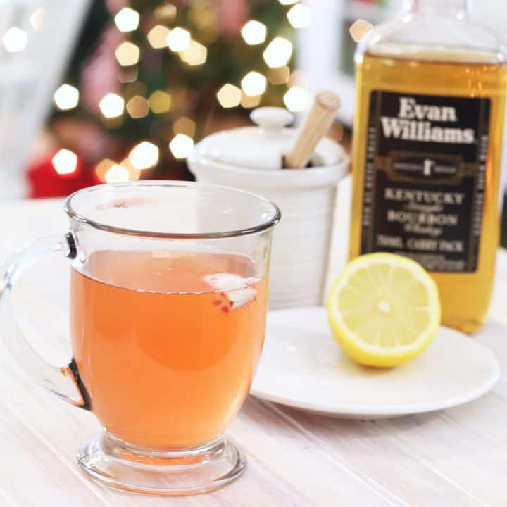 A glass cup with peppermint whiskey honey and lemon on a white tray with a bottle of whiskey and half cut lemon on a plate.